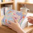 Gros plan sur les mains d'une personne ouvrant un manga coloré sur une table en bois, devant une bibliothèque.